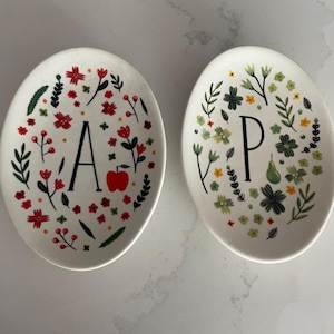 Puede incluir: Dos platos de cer&aacute;mica ovalados. Un plato presenta la letra "A" y una manzana roja, rodeada de dise&ntilde;os florales rojos, verdes y naranjas. El otro plato tiene la letra "P" y dise&ntilde;os florales verdes, amarillos y blancos.