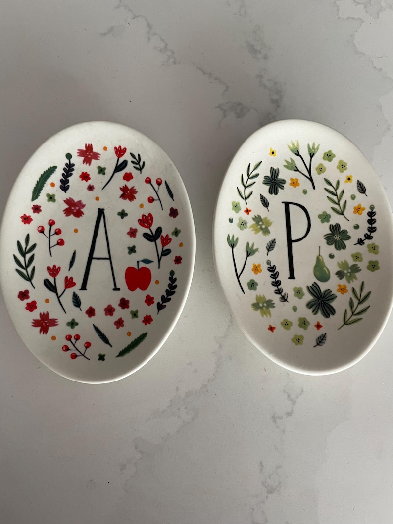 Puede incluir: Dos platos de cer&aacute;mica ovalados. Un plato presenta la letra "A" y una manzana roja, rodeada de dise&ntilde;os florales rojos, verdes y naranjas. El otro plato tiene la letra "P" y dise&ntilde;os florales verdes, amarillos y blancos.