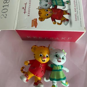 Könnte beinhalten: Ein Hallmark Keepsake Weihnachtsbaumschmuck mit Daniel Tiger und Katerina Kittycat. Daniel Tiger trägt einen roten Kapuzenpullover und Turnschuhe, Katerina ein grünes Kleid. Die Box trägt die Aufschrift "2018" und "Daniel Tiger's Neighborhood".