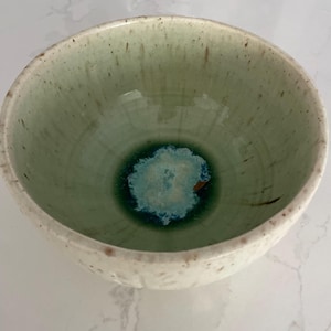 Peut inclure: Un bol en céramique fait à la main avec un intérieur vert clair et un extérieur blanc. L'intérieur du bol présente un centre vert foncé et bleu. Le bord a un aspect texturé et moucheté, ajoutant à son charme rustique.