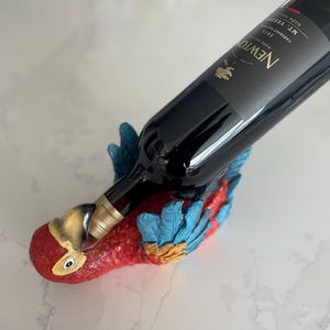 Puede incluir: Un soporte para botellas de vino con forma de loro rojo, azul y amarillo sostiene una botella de vino oscuro. La botella tiene una etiqueta negra con texto blanco, incluyendo "NEWTON". El loro tiene un diseño detallado y texturizado.