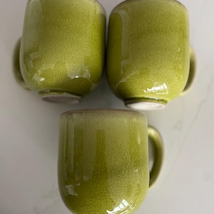 May include: Three green ceramic mugs with a crackle glaze finish. Each mug has a rounded shape, a handle, and a white base. The mugs are arranged in a triangular pattern, showcasing their design and color.