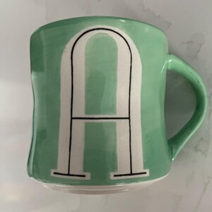 Peut inclure: Une tasse en c&eacute;ramique vert menthe avec la lettre "A" blanche dans une police de caract&egrave;res en gras et soulign&eacute;e. La tasse a une forme incurv&eacute;e et une anse verte assortie. Le design est simple et moderne.