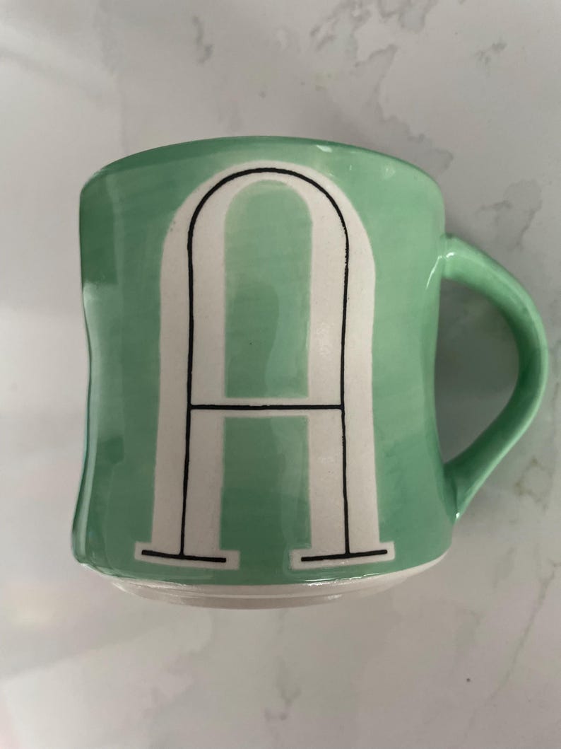 Peut inclure: Une tasse en c&eacute;ramique vert menthe avec la lettre "A" blanche dans une police de caract&egrave;res en gras et soulign&eacute;e. La tasse a une forme incurv&eacute;e et une anse verte assortie. Le design est simple et moderne.