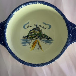 May include: A round, ceramic bowl with a light yellow interior and a dark blue speckled rim. The bowl features a colorful illustration of Mont St. Michel, with the text "Mont St. Michel" in script. The bowl has two handles.