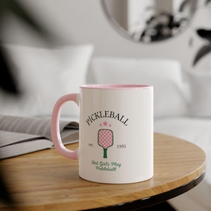Peut inclure: Mug en céramique blanche avec une anse et un intérieur roses. Le mug présente le mot "PICKLEBALL" au-dessus d'un graphique d'une raquette de pickleball et le texte "Hot Girls Play Pickleball".