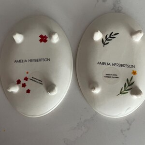 Puede incluir: Dos platos ovalados de cer&aacute;mica blanca con peque&ntilde;os pies. Uno tiene detalles florales rojos, el otro un dise&ntilde;o floral negro y amarillo. El texto "AMELIA HERBERTSON" y "MADE IN CHINA FABRIQUE EN CHINE" est&aacute;n impresos en los platos.