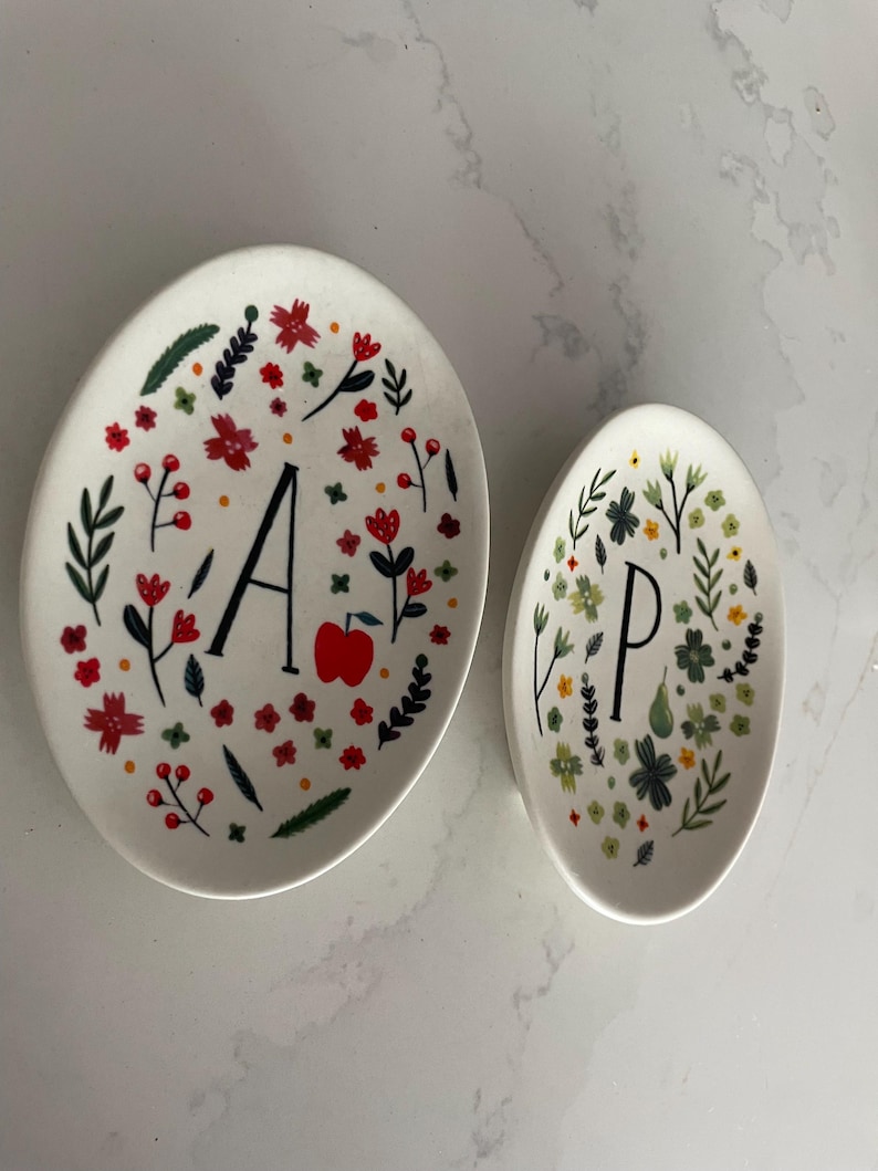 Puede incluir: Dos platos ovalados de cer&aacute;mica blanca. El plato m&aacute;s grande tiene la letra negra "A" y una manzana roja, rodeada de flores rojas y hojas verdes. El plato m&aacute;s peque&ntilde;o tiene la letra negra "P" con dise&ntilde;os florales verdes y amarillos.