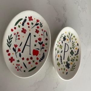 Puede incluir: Dos platos ovalados de cer&aacute;mica blanca. El plato m&aacute;s grande tiene la letra "A" y una manzana roja, rodeada de flores rojas y hojas verdes. El plato m&aacute;s peque&ntilde;o tiene la letra "P" y dise&ntilde;os florales similares.