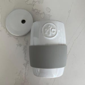 Puede incluir: Un termo de cerámica blanca con una banda de silicona gris y una tapa a juego. El termo tiene un logotipo en la parte delantera. La tapa es redonda con un agujero en el centro. El termo está diseñado para bebidas frías o calientes.