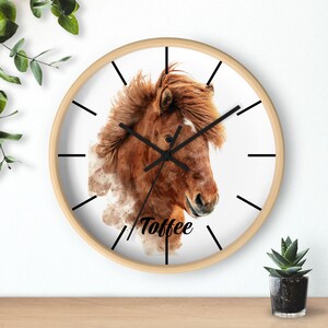 May include: A white clock with a brown horse illustration and the word "Toffee" printed below the horse. The clock has black hands and black markings for the hours and minutes. The clock has a light wood frame.