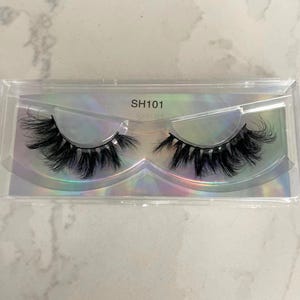 Op de afbeelding: Een paar zwarte valse wimpers in een doorzichtige plastic verpakking. De wimpers zijn lang en vol, met een natuurlijke krul. De verpakking heeft de tekst "SH101" erop.