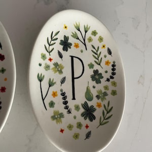 Puede incluir: Plato ovalado de cer&aacute;mica blanca con un dise&ntilde;o floral y la letra "P". El dise&ntilde;o incluye flores verdes, amarillas y naranjas, hojas verdes y una pera verde. La letra "P" es negra.