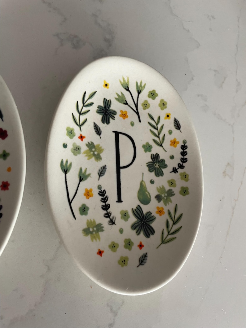 Puede incluir: Plato ovalado de cer&aacute;mica blanca con un dise&ntilde;o floral y la letra "P". El dise&ntilde;o incluye flores verdes, amarillas y naranjas, hojas verdes y una pera verde. La letra "P" es negra.