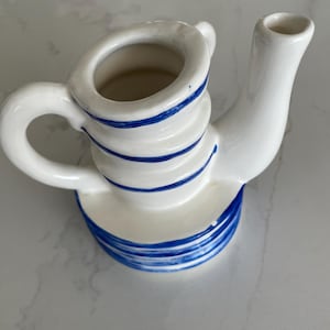 Puede incluir: Cremera de cerámica blanca con rayas azules. La cremera tiene un asa, un pico y una base circular. El diseño presenta rayas azules horizontales alrededor del cuerpo y la base, creando una estética clásica y náutica.