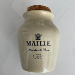 Puede incluir: Un tarro de mostaza de cerámica de color crema con un tapón de corcho. El tarro presenta el texto "MAILLE. Moutarde Fine" en escritura negra, con "DEPUIS 1747" debajo. El tarro tiene un diseño clásico y vintage.