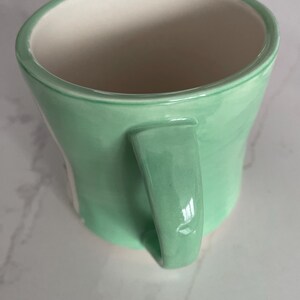 Peut inclure: Une tasse en c&eacute;ramique vert clair avec une forme incurv&eacute;e et une anse assortie. L'int&eacute;rieur de la tasse est blanc. La tasse est con&ccedil;ue pour contenir des boissons chaudes ou froides.