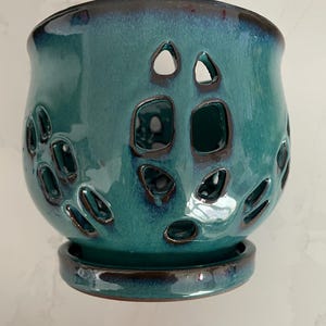 May include: A teal ceramic planter with a glossy finish and a dark brown rim. The planter features decorative cut-out holes and sits on a matching saucer. The design is suitable for small plants or succulents.