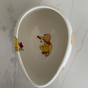 May include: White ceramic bowl with a whimsical illustration of a baby in yellow plaid pants and a red shirt. The bowl has a unique, elongated shape and is decorated with red and yellow accents.