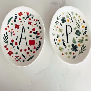 Puede incluir: Dos platos decorativos blancos de forma ovalada. Un plato presenta la letra "A" y una manzana roja, rodeada de dise&ntilde;os florales rojos y verdes. El otro plato tiene la letra "P" y una pera, con dise&ntilde;os florales verdes, amarillos y blancos.