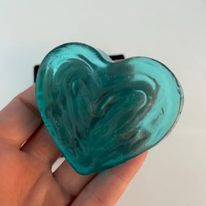 Puede incluir: Un objeto en forma de corazón de color turquesa con un aspecto veteado. El material translúcido tiene remolinos oscuros. El corazón se sostiene en una mano sobre un fondo blanco. El objeto es probablemente de vidrio.