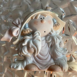 Könnte beinhalten: Keramikfigur eines sitzenden Kindes mit blonden Haaren, Sommersprossen und einem Hut mit rosa Schleife. Das Kind trägt ein weißes Kleid und hat einen flauschigen, fellartigen Besatz. Die Figur sitzt.