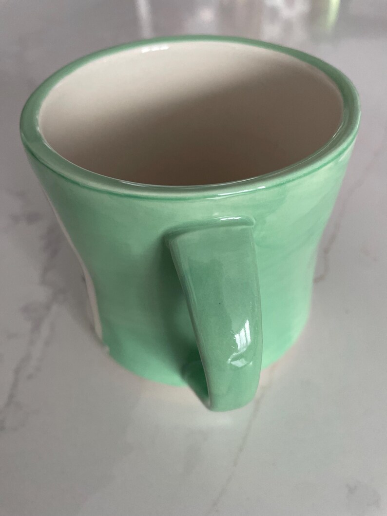 Peut inclure: Une tasse en c&eacute;ramique vert clair avec une forme incurv&eacute;e et une anse assortie. L'int&eacute;rieur de la tasse est blanc, contrastant avec l'ext&eacute;rieur. La tasse est con&ccedil;ue pour contenir des boissons chaudes ou froides.