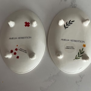 Puede incluir: Dos platos ovalados de cer&aacute;mica blanca con peque&ntilde;os pies. Uno presenta dise&ntilde;os florales rojos, el otro tiene un dise&ntilde;o floral verde y amarillo. Ambos platos tienen el texto "AMELIA HERBERTSON" y "MADE IN CHINA FABRIQUE EN CHINE".