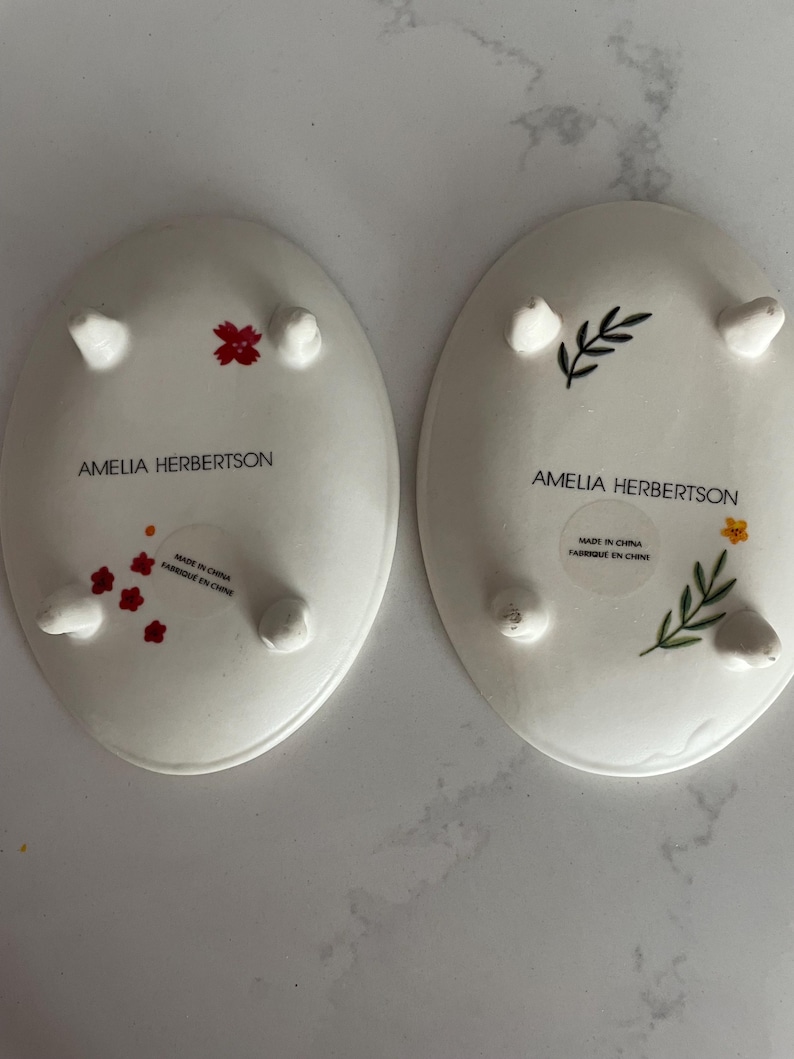 Puede incluir: Dos platos ovalados de cer&aacute;mica blanca con peque&ntilde;os pies. Uno presenta dise&ntilde;os florales rojos, el otro tiene un dise&ntilde;o floral verde y amarillo. Ambos platos tienen el texto "AMELIA HERBERTSON" y "MADE IN CHINA FABRIQUE EN CHINE".