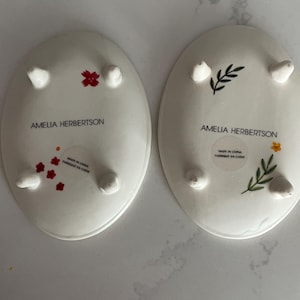 Puede incluir: Dos platos ovalados de cer&aacute;mica blanca con peque&ntilde;os pies. Uno tiene detalles florales rojos, el otro detalles florales verdes y amarillos. El texto "AMELIA HERBERTSON" y "MADE IN CHINA FABRIQUE EN CHINE" est&aacute; impreso en la parte inferior de cada plato.
