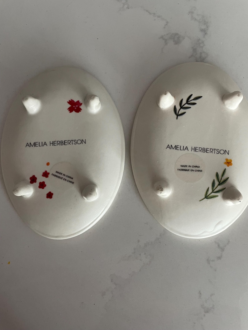 Puede incluir: Dos platos ovalados de cer&aacute;mica blanca con peque&ntilde;os pies. Uno tiene detalles florales rojos, el otro detalles florales verdes y amarillos. El texto "AMELIA HERBERTSON" y "MADE IN CHINA FABRIQUE EN CHINE" est&aacute; impreso en la parte inferior de cada plato.
