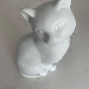 May include: A small, white ceramic cat figurine in a seated position. The cat has pointed ears, a smooth, glossy finish, and is looking down. The figurine is a decorative item, likely for display on a shelf or table.