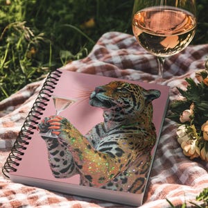 May include: A pink notebook with a spiral binding, depicting a jaguar with a martini. The jaguar has glitter and orange nail polish. A glass of rosé wine is next to the notebook on a checkered blanket, with flowers and grass in the background.