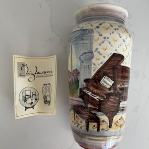 May include: A hand-painted earthenware vase with a piano design. The vase features a brown grand piano with sheet music, set against a backdrop of curtains and a patterned wall. The vase is labeled "Liz Lawrence, painted earthenware."