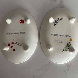 Puede incluir: Dos platos ovalados de cer&aacute;mica blanca con dise&ntilde;os florales y peque&ntilde;os pies. Uno presenta flores rojas, el otro una flor amarilla y hojas verdes. El texto "AMELIA HERBERTSON" y "MADE IN CHINA FABRIQUE EN CHINE" est&aacute; impreso en los platos.