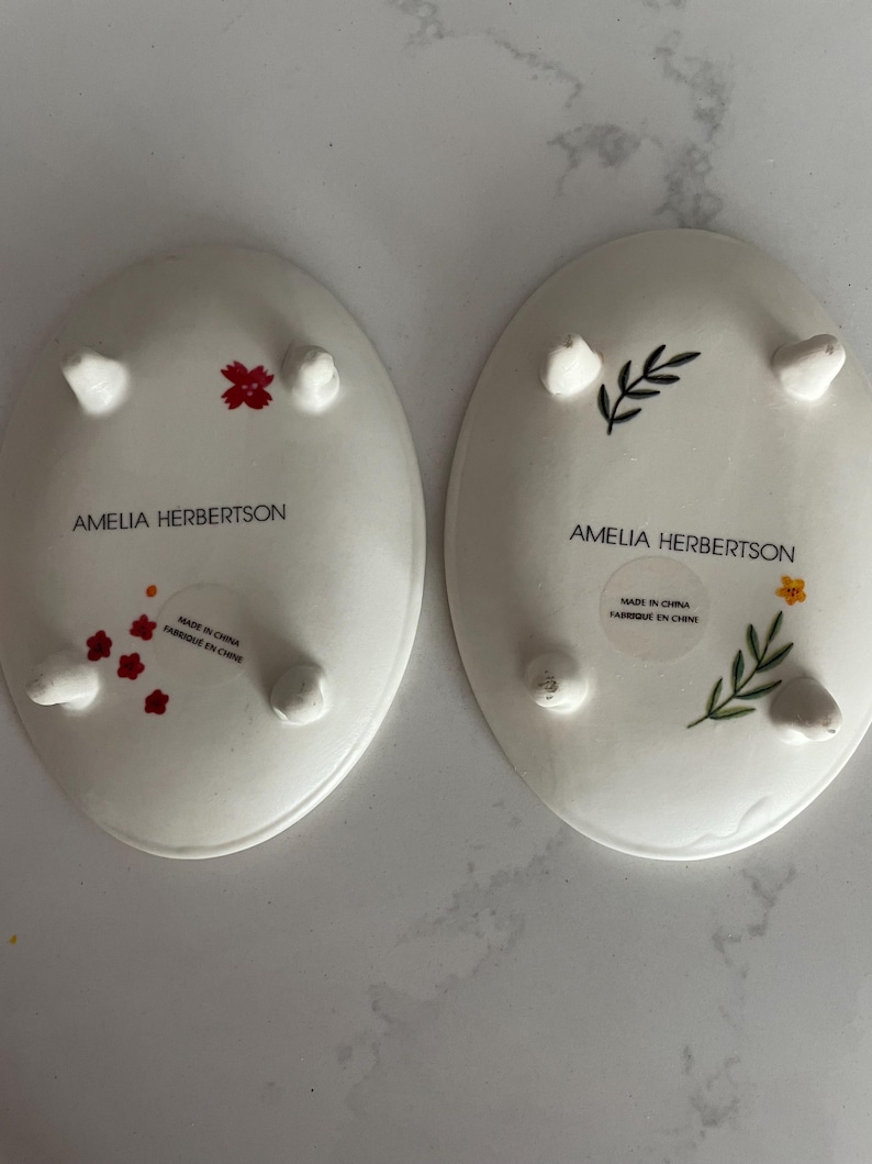 Puede incluir: Dos platos ovalados de cer&aacute;mica blanca con dise&ntilde;os florales y peque&ntilde;os pies. Uno presenta flores rojas, el otro una flor amarilla y hojas verdes. El texto "AMELIA HERBERTSON" y "MADE IN CHINA FABRIQUE EN CHINE" est&aacute; impreso en los platos.