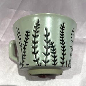 May include: A light green ceramic teacup with a small handle. The cup features a black floral design. The cup is approximately 3 inches tall and 3 inches wide.