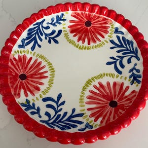 May include: A round, white ceramic plate with a red scalloped edge. The plate features a floral design with red flowers, blue leaves, and green accents. The plate is a decorative piece for serving food.