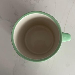 Peut inclure: Une tasse en c&eacute;ramique avec un bord et une anse vert clair. L'int&eacute;rieur de la tasse est de couleur beige clair. La tasse est vide et semble propre.