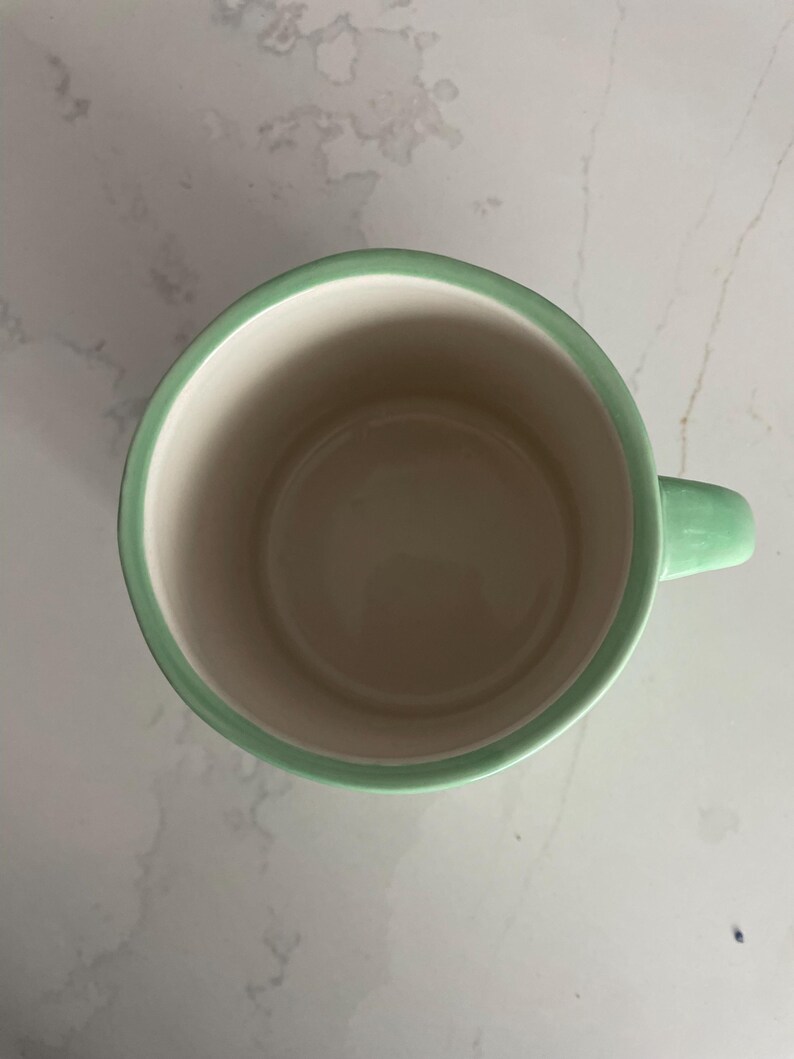 Peut inclure: Une tasse en c&eacute;ramique avec un bord et une anse vert clair. L'int&eacute;rieur de la tasse est de couleur beige clair. La tasse est vide et semble propre.
