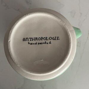 Peut inclure: Une tasse en c&eacute;ramique blanche avec une anse et un bord vert clair. Le fond de la tasse porte l'inscription "ANTHROPOLOGIE hand painted" en noir. La tasse est pos&eacute;e sur un fond blanc et gris.