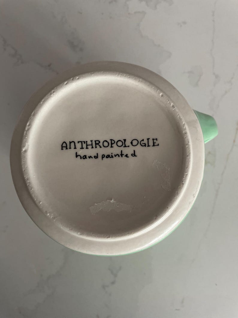 Peut inclure: Une tasse en c&eacute;ramique blanche avec une anse et un bord vert clair. Le fond de la tasse porte l'inscription "ANTHROPOLOGIE hand painted" en noir. La tasse est pos&eacute;e sur un fond blanc et gris.