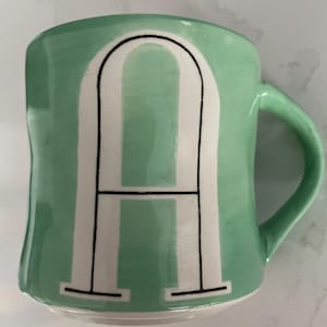 Peut inclure: Une tasse en c&eacute;ramique vert menthe avec la lettre "A" blanche en style bloc. La lettre est soulign&eacute;e en noir. La tasse a une anse incurv&eacute;e et une finition brillante.