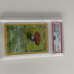 Puede incluir: Una carta de Pok&eacute;mon calificada con Vileplume del set Jungle de 1999. La carta est&aacute; dentro de un soporte protector transparente y est&aacute; calificada NM-MT 8. La ilustraci&oacute;n de la carta representa una criatura con forma de hongo rojo y morado.