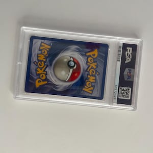 Puede incluir: Una carta Pok&eacute;mon calificada, encapsulada en un soporte de pl&aacute;stico transparente. La carta presenta el logotipo de Pok&eacute;mon y un dise&ntilde;o de Pok&eacute; Ball sobre un fondo azul. El soporte tiene una etiqueta PSA con c&oacute;digo de barras y n&uacute;mero de serie.