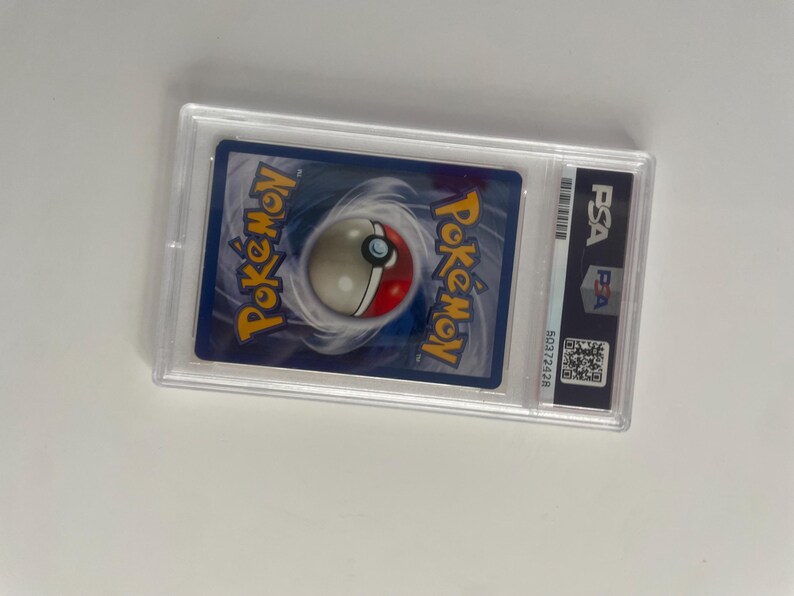 Puede incluir: Una carta Pok&eacute;mon calificada, encapsulada en un soporte de pl&aacute;stico transparente. La carta presenta el logotipo de Pok&eacute;mon y un dise&ntilde;o de Pok&eacute; Ball sobre un fondo azul. El soporte tiene una etiqueta PSA con c&oacute;digo de barras y n&uacute;mero de serie.