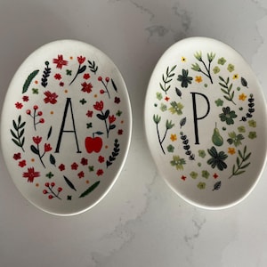 Puede incluir: Dos platos de cer&aacute;mica ovalados blancos. Uno presenta la letra "A" rodeada de flores rojas, hojas verdes y una manzana roja. El otro tiene la letra "P" con dise&ntilde;os florales verdes y amarillos.