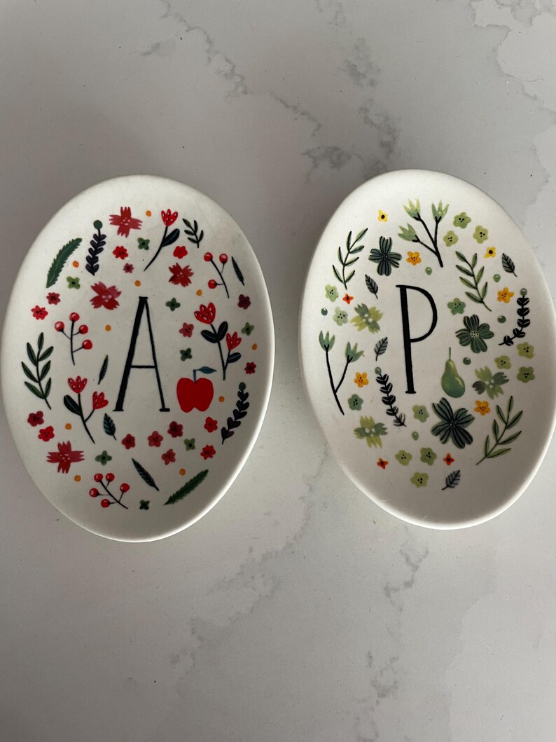 Puede incluir: Dos platos de cer&aacute;mica ovalados blancos. Uno presenta la letra "A" rodeada de flores rojas, hojas verdes y una manzana roja. El otro tiene la letra "P" con dise&ntilde;os florales verdes y amarillos.