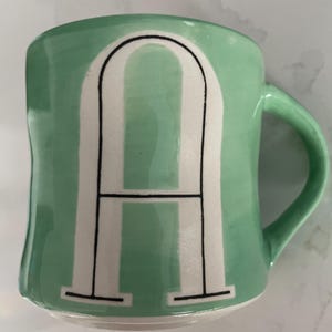 Peut inclure: Une tasse en c&eacute;ramique vert menthe avec la lettre "A" blanche en caract&egrave;res gras, soulign&eacute;e de noir. La tasse a une anse incurv&eacute;e et une forme l&eacute;g&egrave;rement irr&eacute;guli&egrave;re. Id&eacute;ale pour le caf&eacute; ou le th&eacute;.