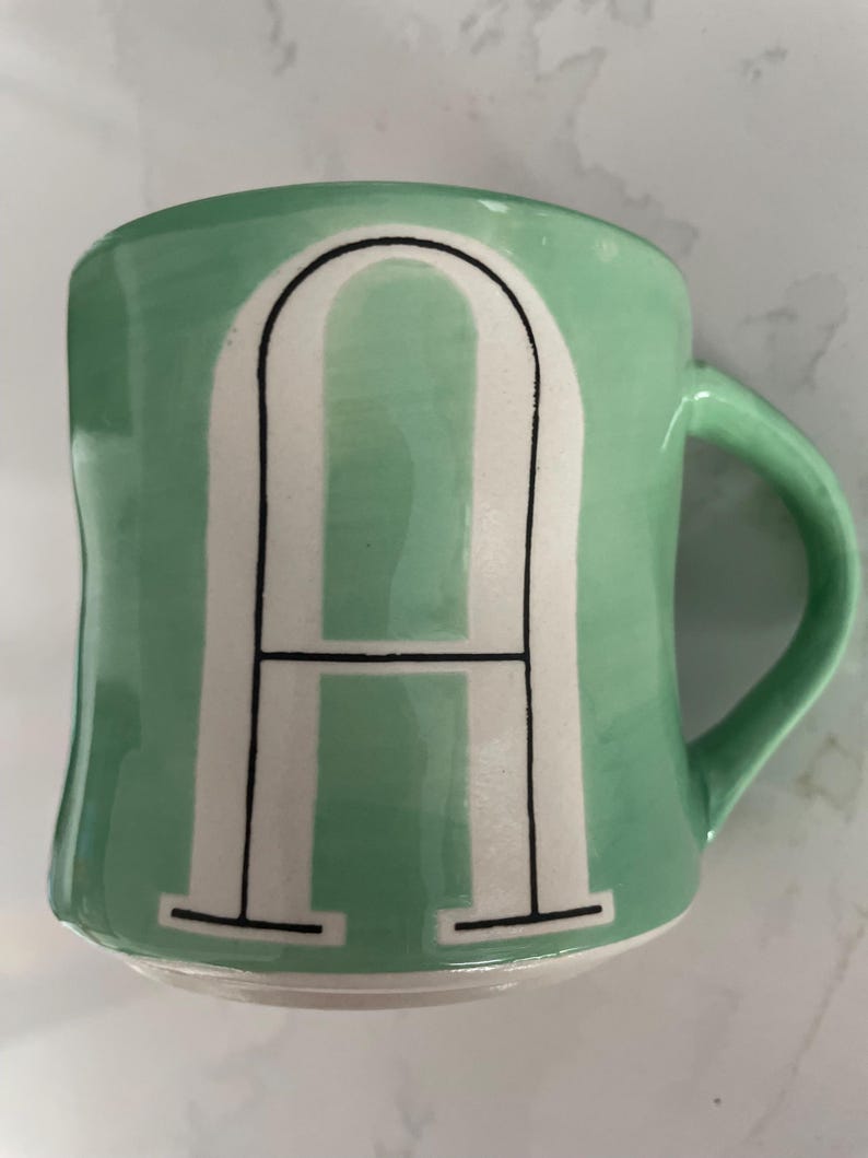Peut inclure: Une tasse en c&eacute;ramique vert menthe avec la lettre "A" blanche en caract&egrave;res gras, soulign&eacute;e de noir. La tasse a une anse incurv&eacute;e et une forme l&eacute;g&egrave;rement irr&eacute;guli&egrave;re. Id&eacute;ale pour le caf&eacute; ou le th&eacute;.